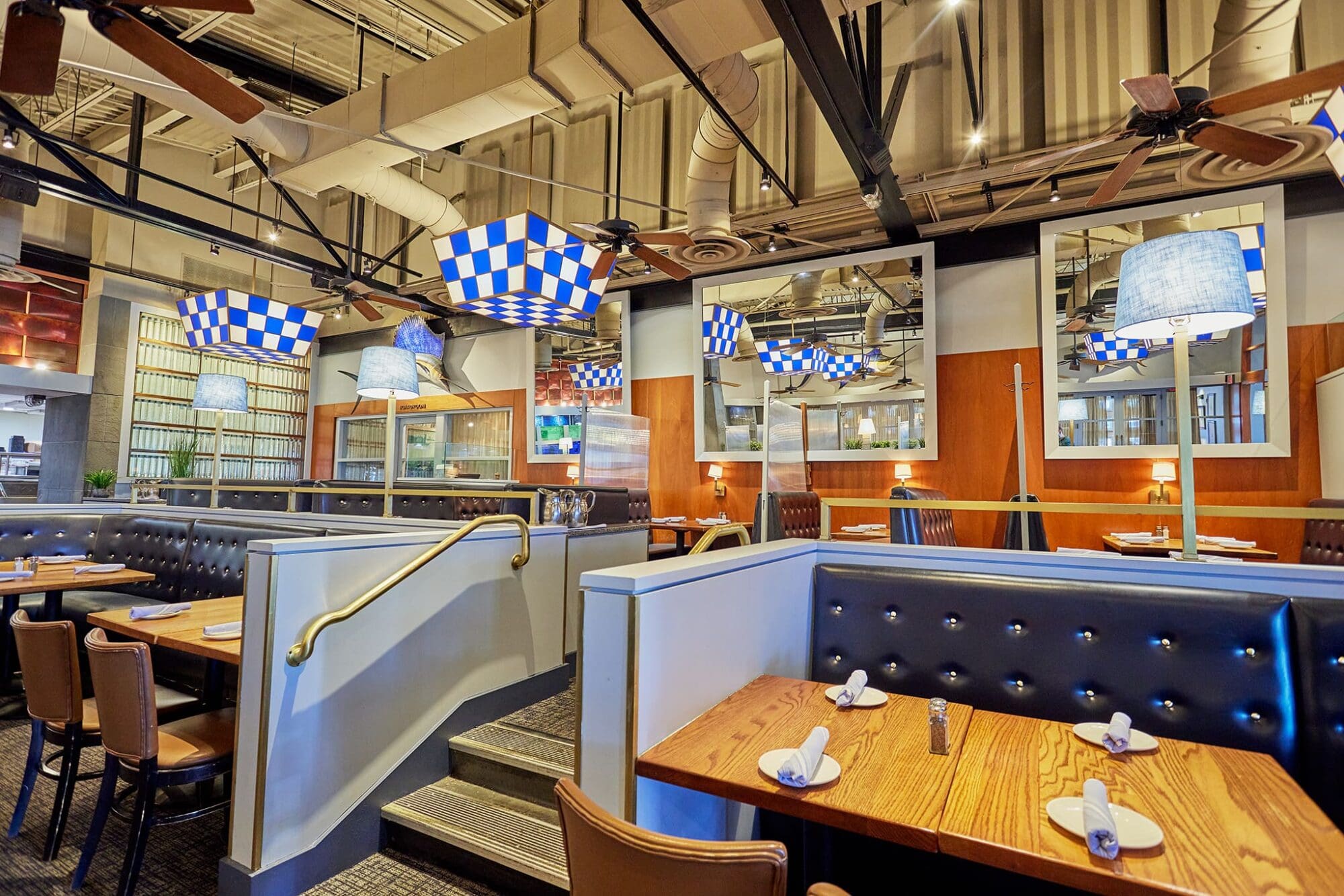 Interior of a modern restaurant with wooden tables, black cushioned booths, and checkered blue and white ceiling lights. The space is well-lit with large mirrors, industrial-style ceiling, and neatly set tables with napkins and salt shakers.