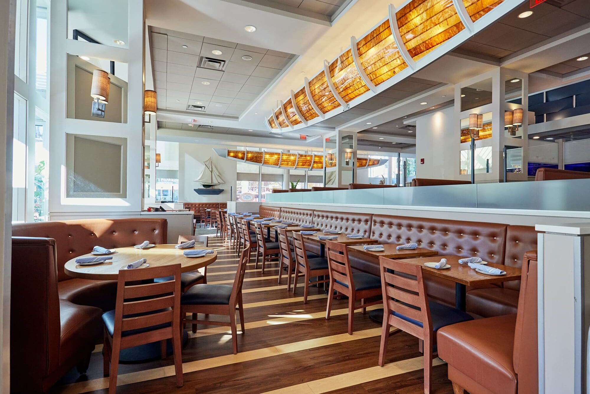 A bright, modern restaurant interior with wooden tables, cushioned brown booths, and chairs. Large windows allow natural light in. Nautical decor includes a ship model. Soft overhead lights and white walls create a welcoming atmosphere.