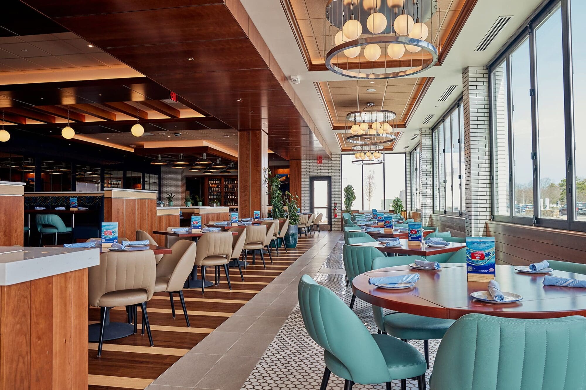 Modern restaurant interior with wooden accents and large windows providing natural light. Tables set with blue napkins and tan and teal chairs. Ceiling features hanging round lights. Floors have a mix of wood and tile patterns.