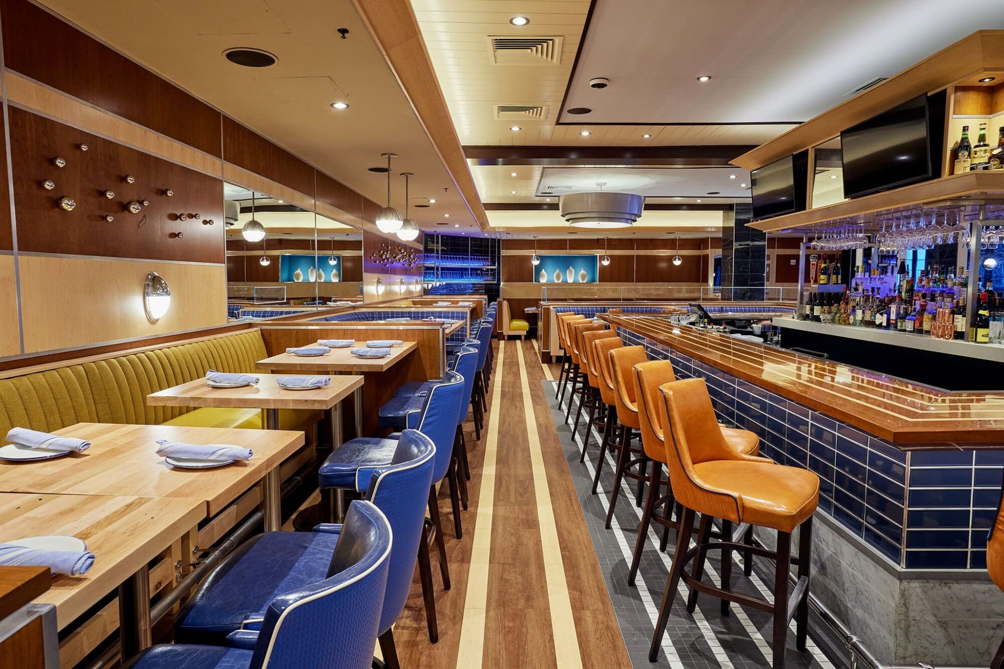 Interior of a modern restaurant featuring wooden tables, blue and orange leather chairs, and a long bar with shelves of bottles. The lighting is warm, and the space has a mix of wooden and mirrored walls, creating a cozy atmosphere.