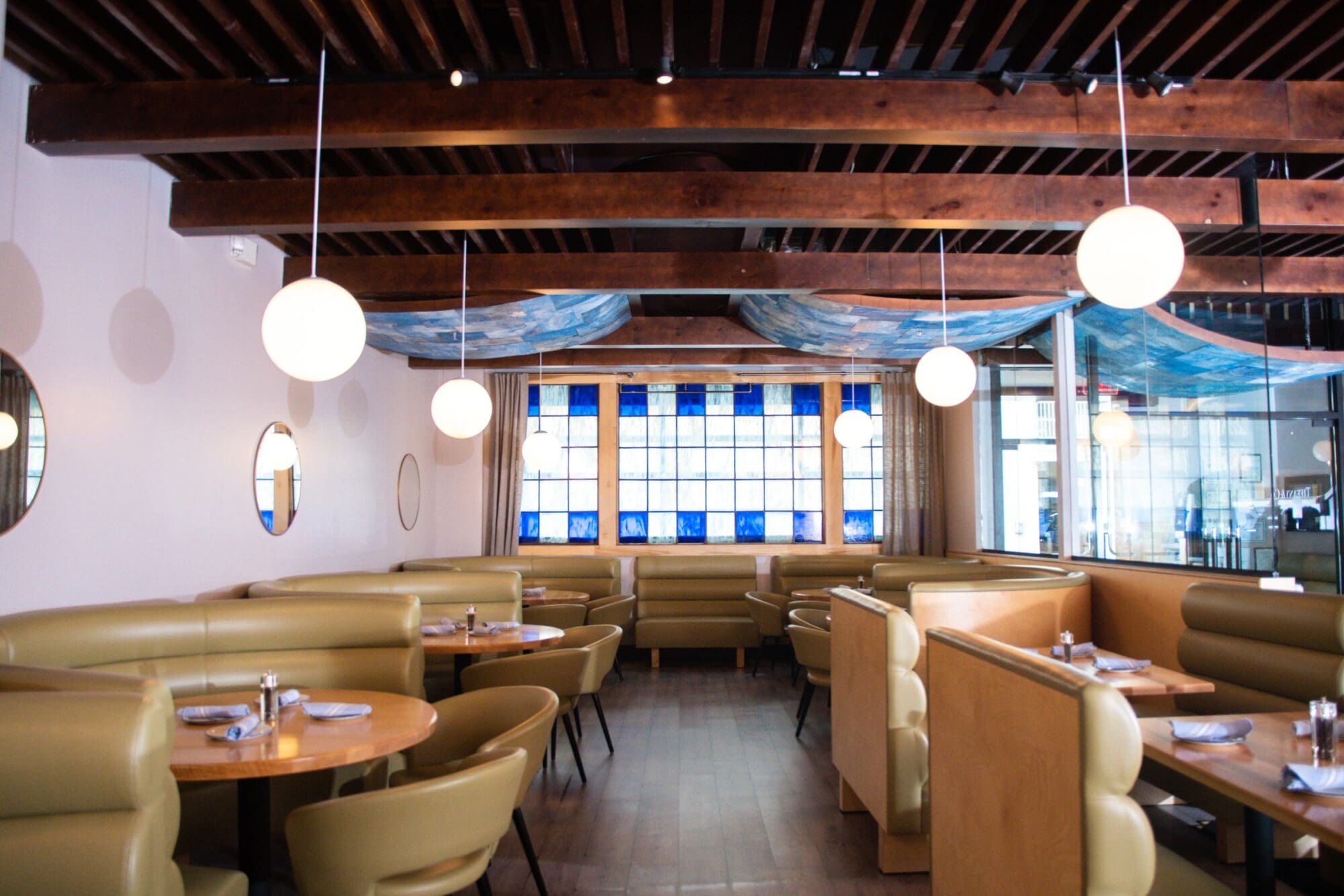 A cozy restaurant interior with wooden ceiling beams, round hanging lights, and circular mirrors on the walls. The space features tan booth seating around circular wooden tables, set with plates and silverware. A stained glass window is visible in the background.