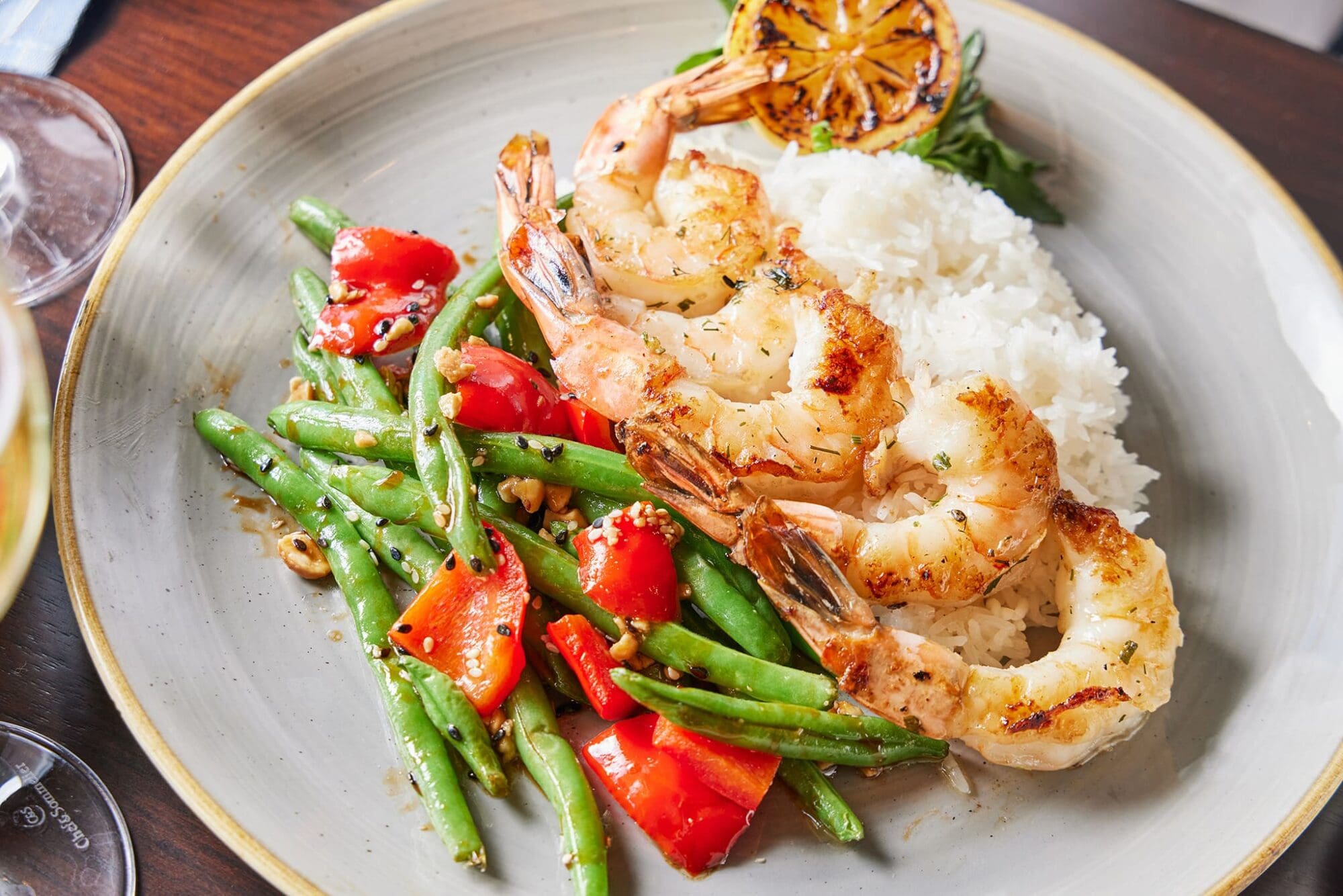 Grilled shrimp skewers served with white rice and a side of green beans and red bell peppers, garnished with sesame seeds. A charred lemon slice is placed on top, all on a round gray plate.