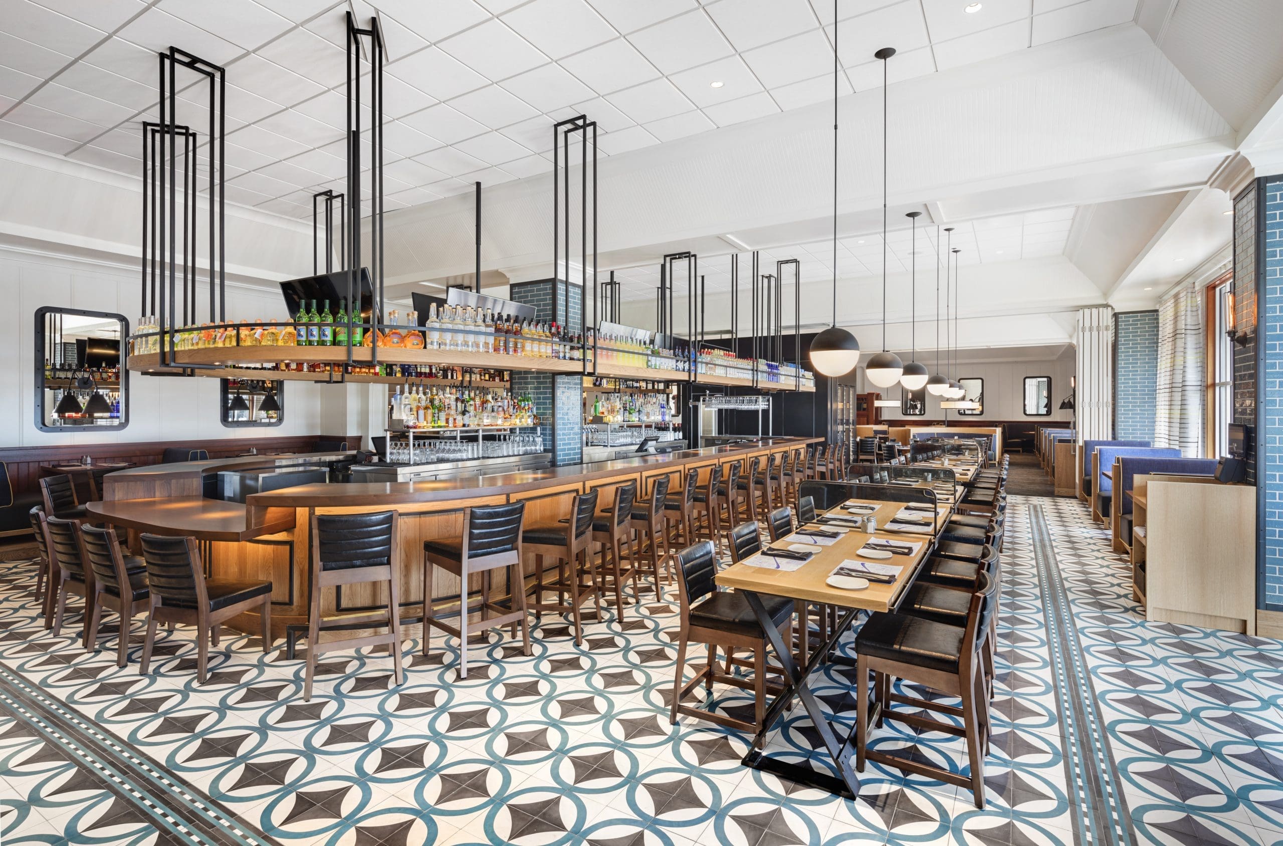 Modern restaurant interior with a central bar surrounded by high wooden chairs. Tables with place settings are arranged in a geometric-tiled floor area. Pendant lights and large windows create a bright, welcoming atmosphere.