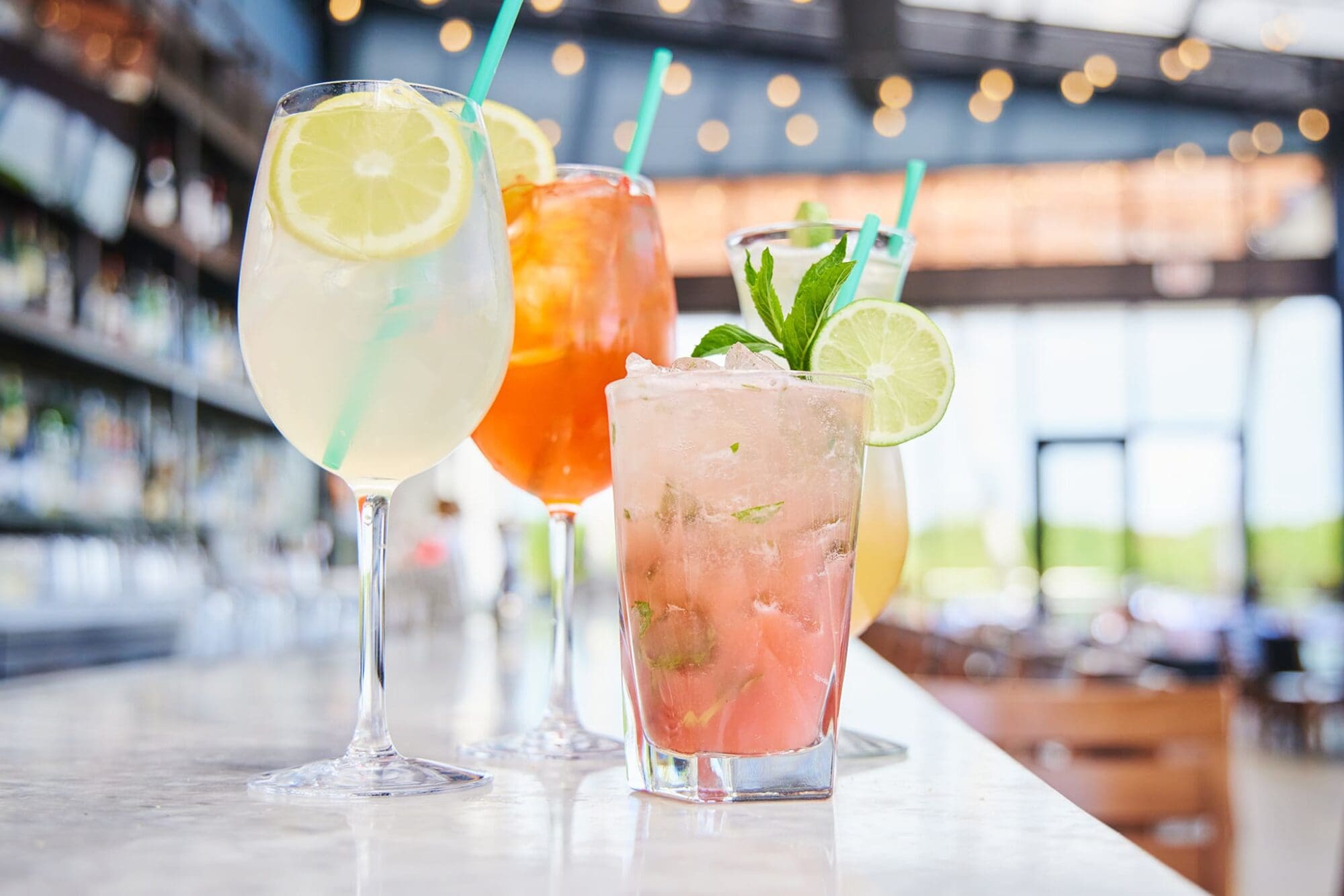 A row of colorful cocktails on a marble bar counter with blurred background. Drinks include a lemon slice garnished glass, orange beverage, and a pink drink with mint and lime garnish. A brightly lit bar with string lights is in the background.