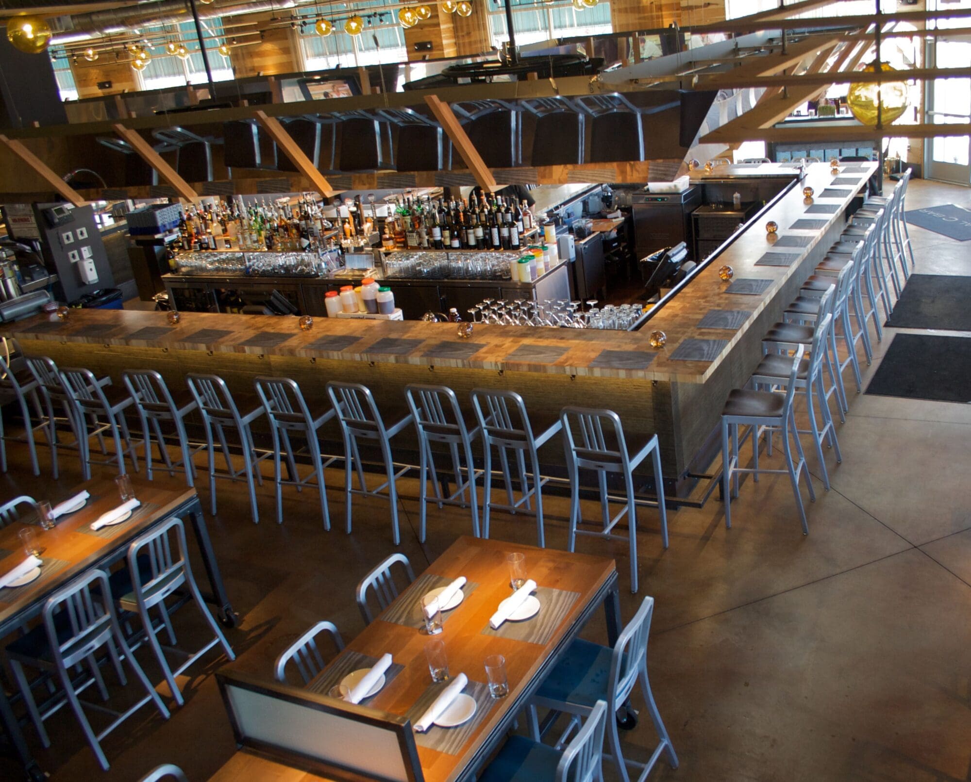 A restaurant with a long, L-shaped bar surrounded by high metal stools. The bar is stocked with various bottles. In the foreground, there are wooden tables set with white napkins and silverware. Large windows provide natural light.