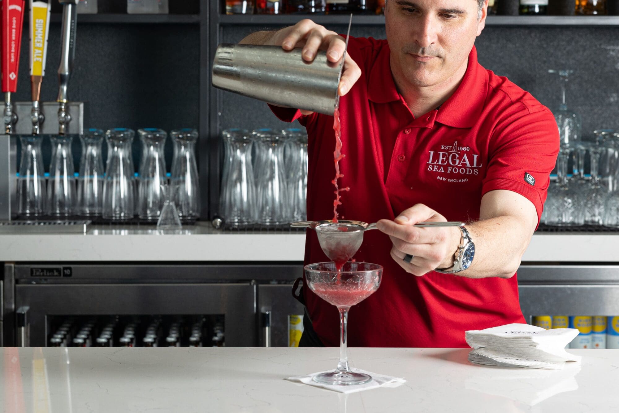 A bartender in a red shirt pours a pink cocktail from a shaker through a strainer into a glass on a bar counter. The background features shelves with glassware and beverage faucets.