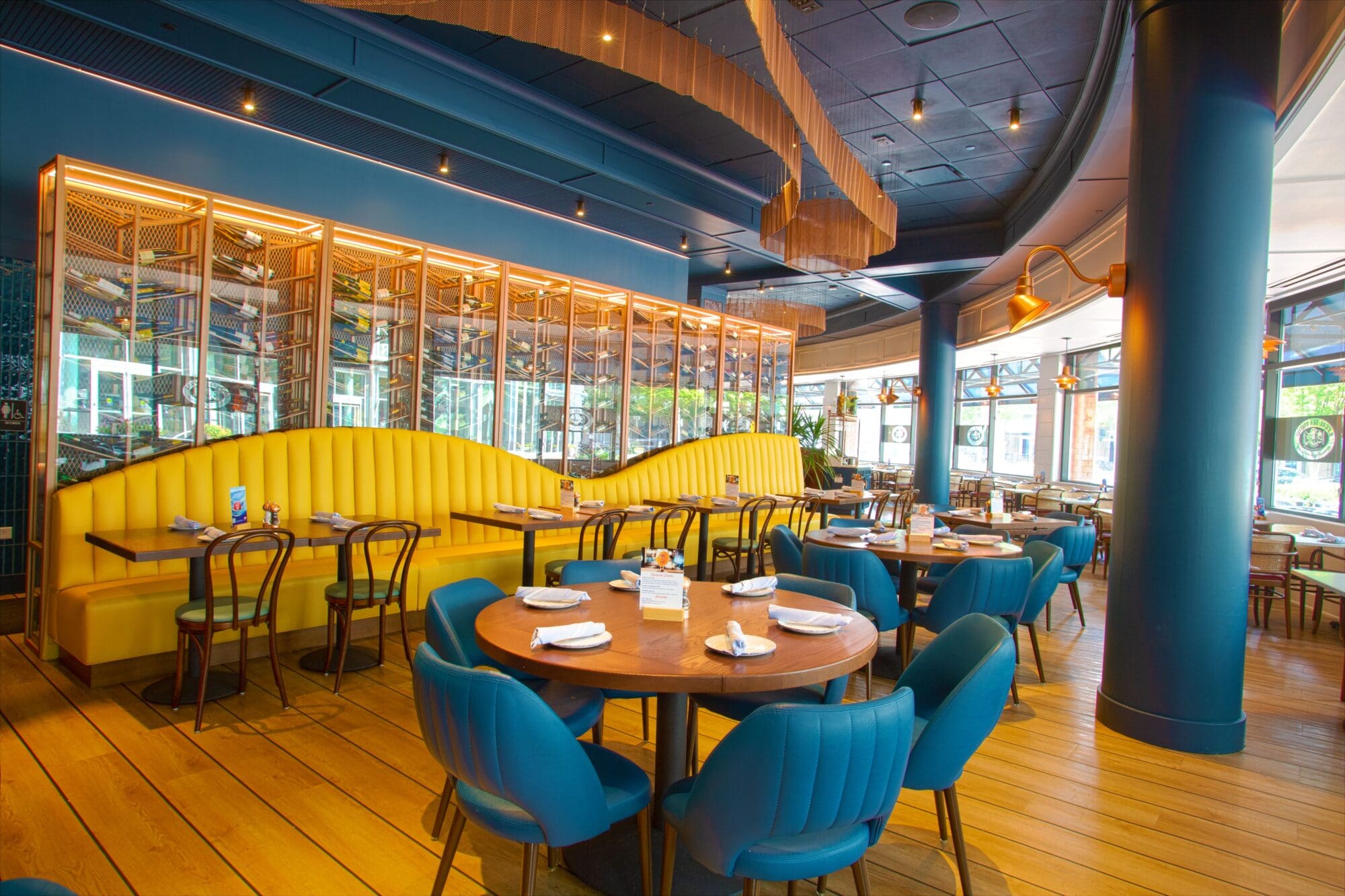 A modern restaurant interior with blue and yellow accents. Curved yellow booth seating lines the wall, with round wooden tables and blue upholstered chairs. The wooden floor and large windows create a bright, inviting atmosphere.