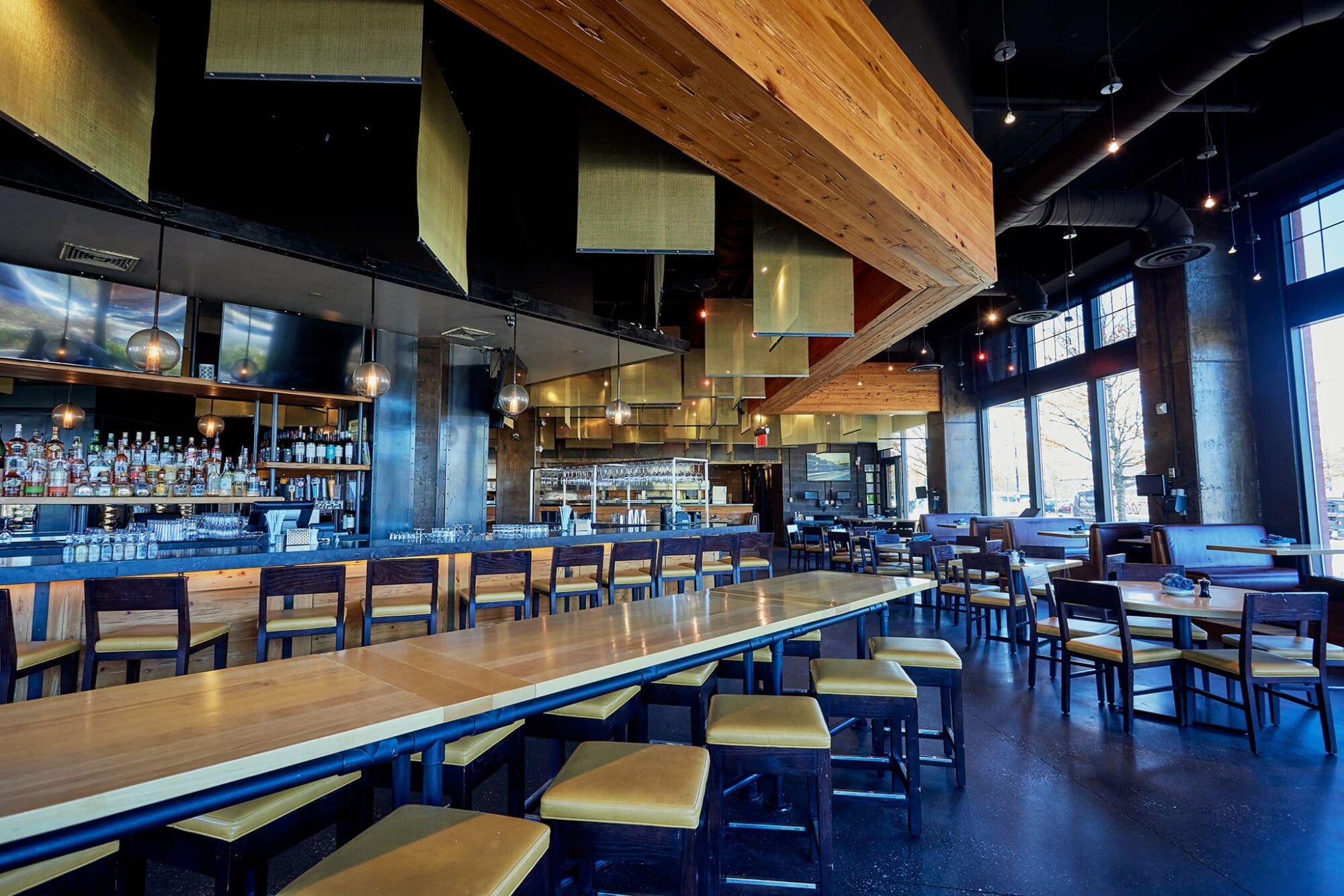 Modern restaurant interior with high ceilings, featuring long wooden tables and chairs. The space has a sleek bar area with shelves of bottles, pendant lighting, and metallic accents. Large windows allow natural light to illuminate the dining area.