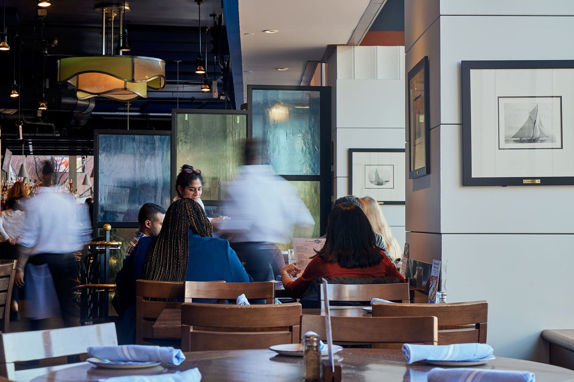 Restaurant interior with a group of people sitting at a table, engaged in conversation. Servers are moving around, creating a blurred effect. Modern decor with framed artwork on the walls, wooden tables, and neatly arranged napkins.