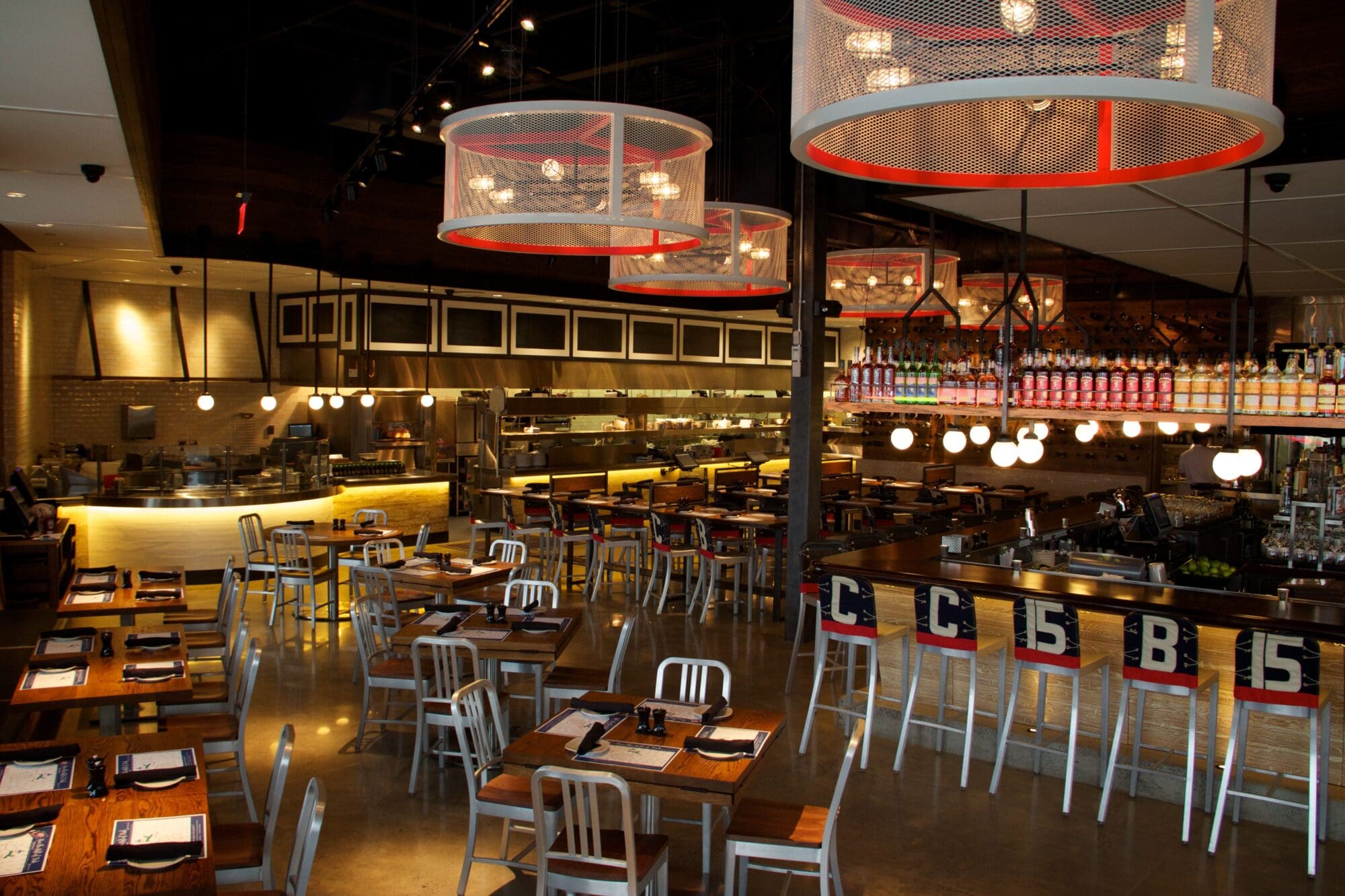 A modern restaurant interior with metal chairs and wooden tables set for service. Large circular light fixtures and a well-stocked bar with hanging glasses are visible. The space has an open kitchen and a warm, inviting ambiance.