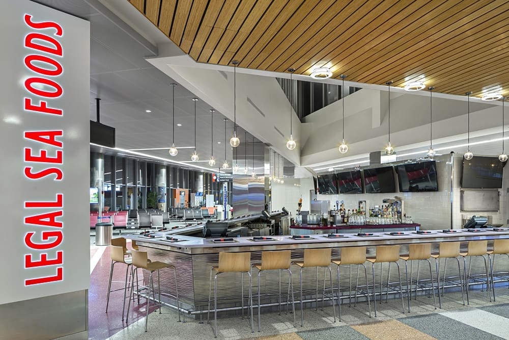 A modern seafood restaurant interior with a sleek bar counter surrounded by wooden stools. The ceiling features wooden panels and hanging lights. A large sign reads 