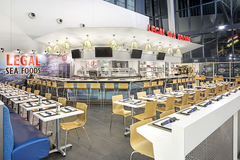 Modern seafood restaurant interior with light wood chairs and tables set with blue place settings. A central bar area and open kitchen are visible under branded signage. Contemporary decor is highlighted by large windows and bright lighting.