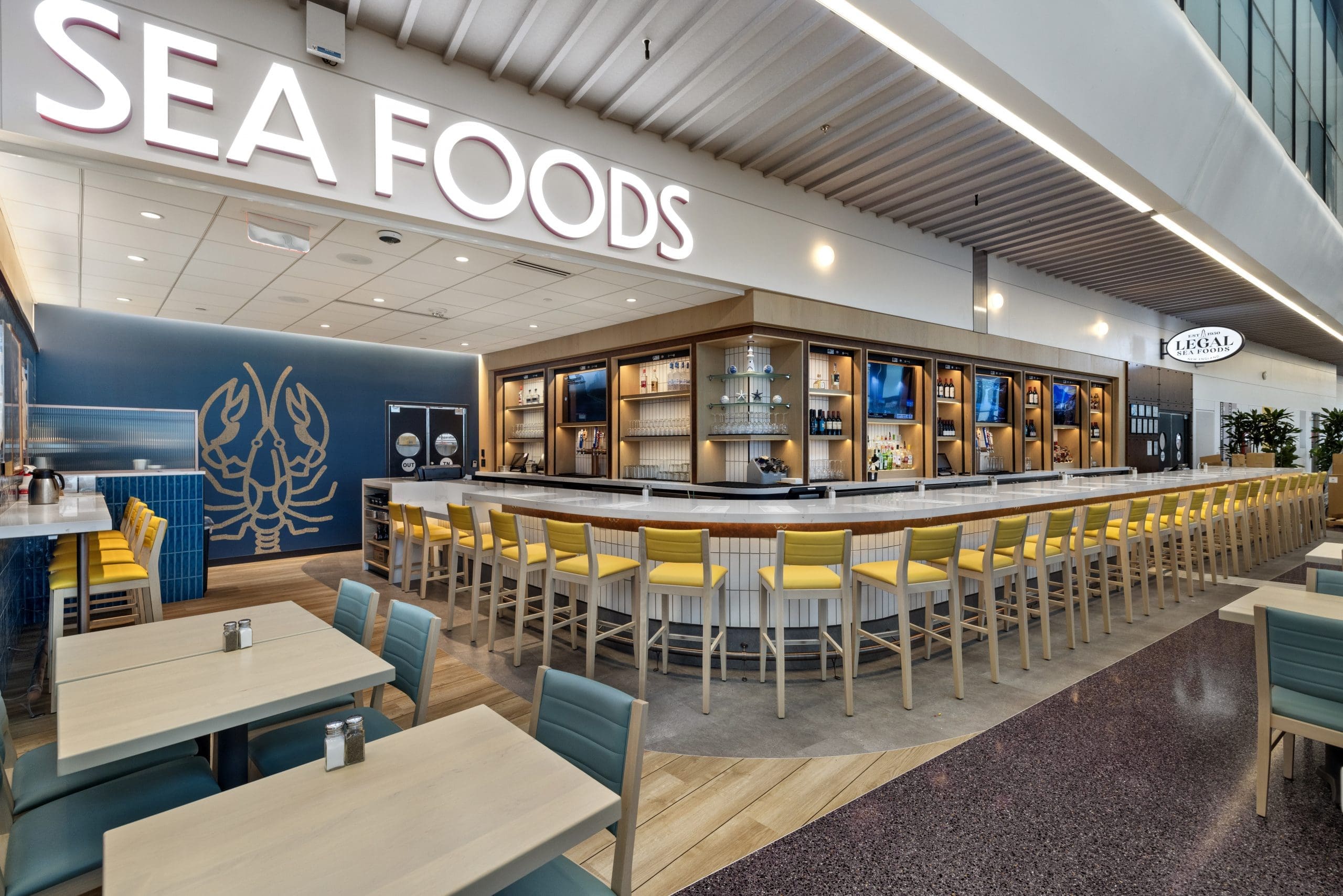 A modern seafood restaurant with a large, curved bar surrounded by yellow chairs. The wall displays shelves of glassware and bottles. 