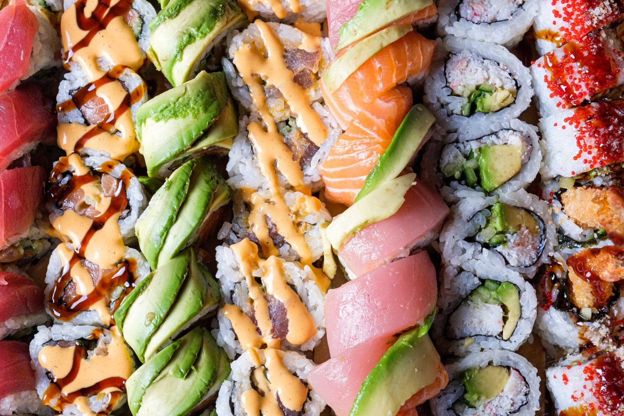 Close-up of an assortment of sushi rolls, including pieces topped with avocado, salmon, and tuna. Some rolls are drizzled with sauces, while others are garnished with red tobiko. The colorful display showcases various textures and flavors.