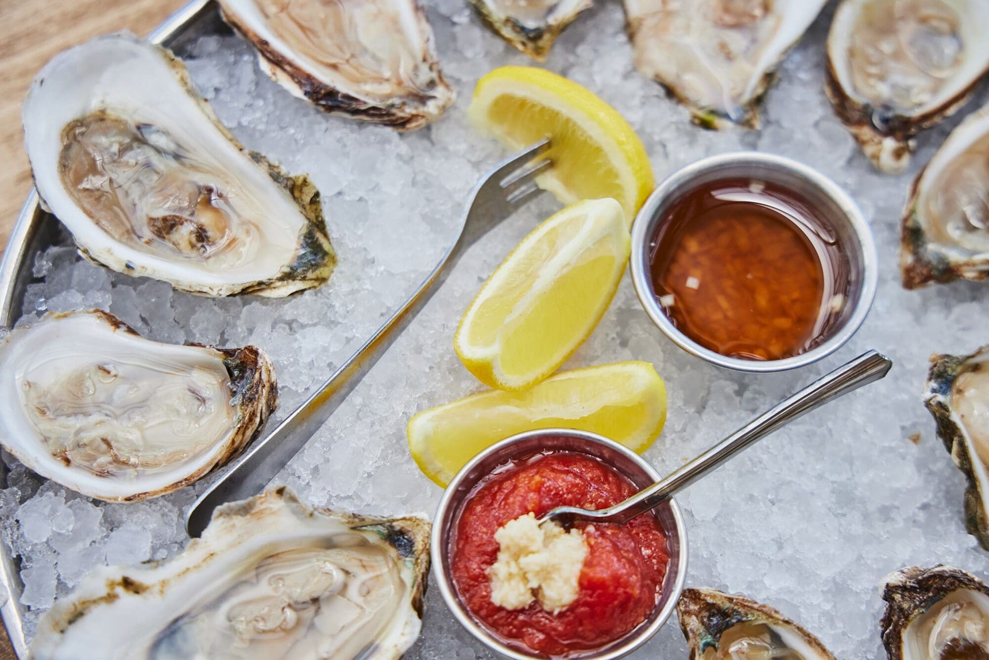 A tray of half-shell oysters on crushed ice, served with lemon wedges, a red cocktail sauce topped with horseradish, and a brown sauce in metal cups. Two small forks are placed on the tray.