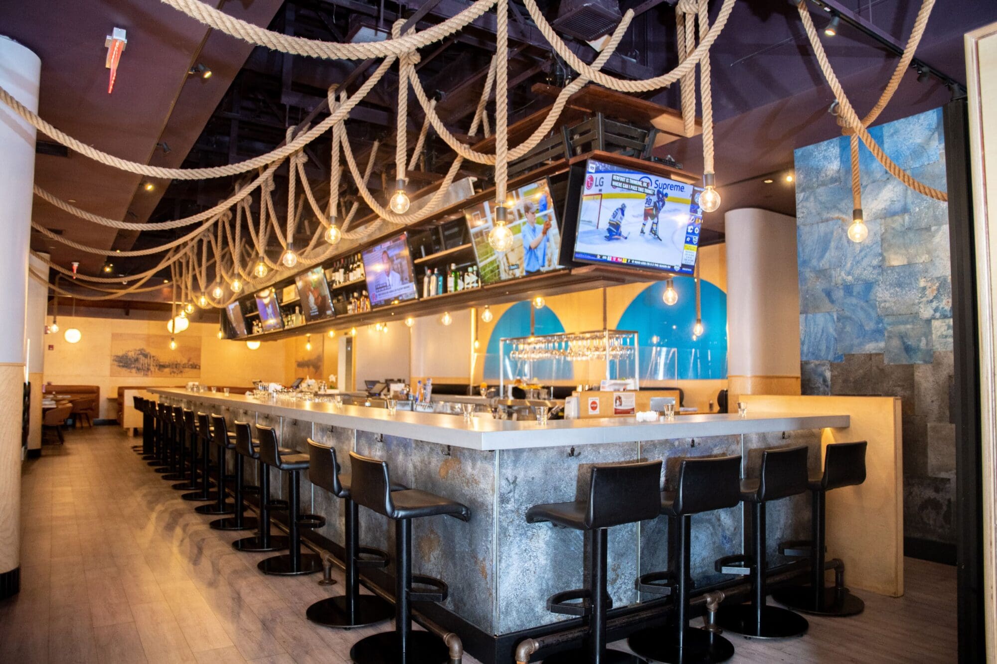 A modern bar with a long counter lined with black stools, overhead rope decorations, and multiple screens showing sports. Pendant lights hang from the ceiling, creating a warm ambiance.