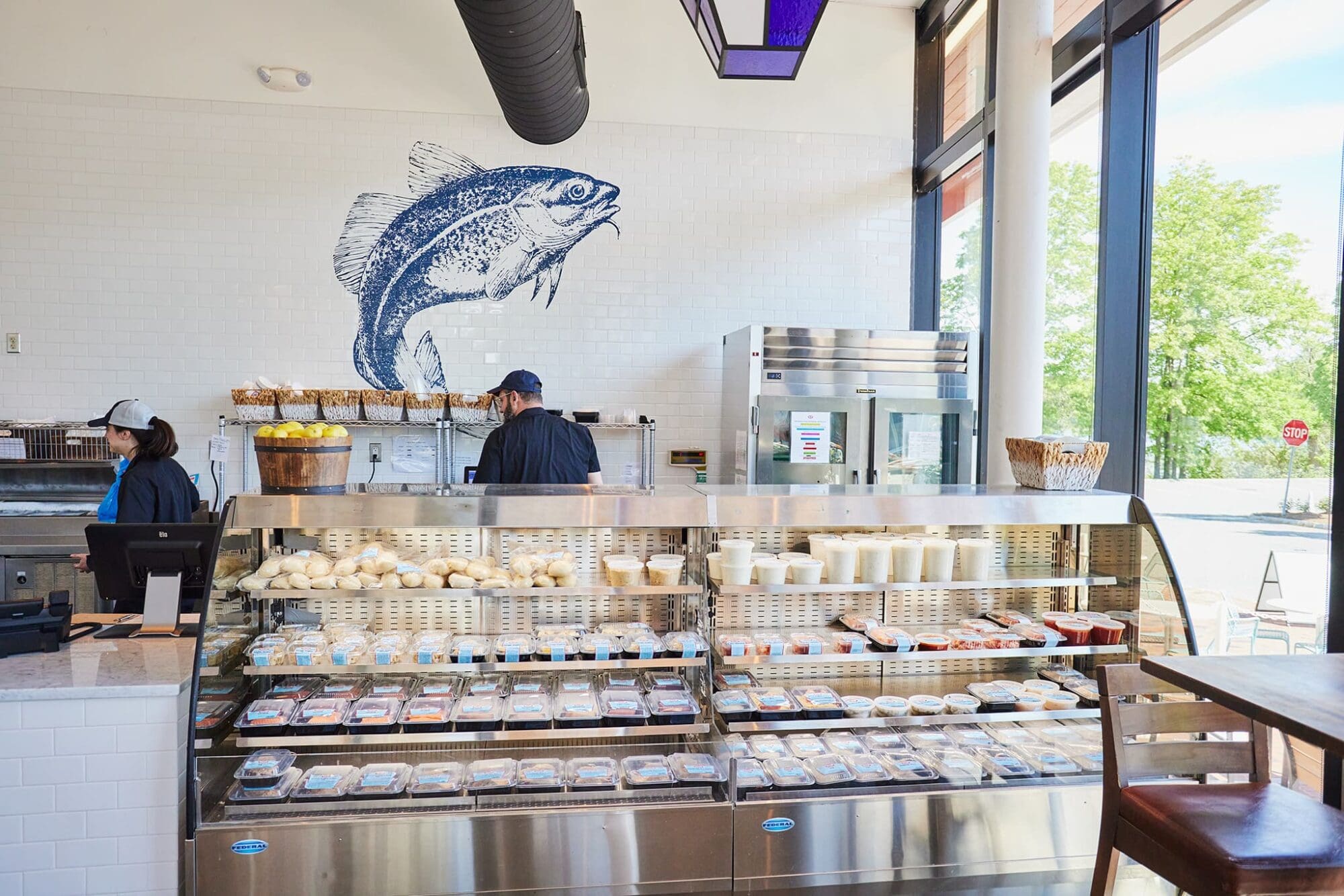 A bright, modern seafood shop interior with a large fish mural on the wall. Two workers are visible behind the counter, which is filled with seafood products. The shop has large windows letting in natural light.