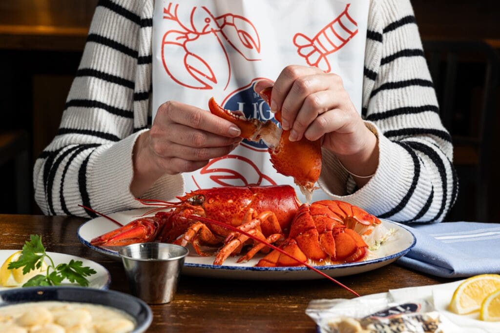 Person in striped sweater cracking open a lobster claw at a restaurant. A white bib with red seafood designs is visible. Plate includes a whole lobster, lemon wedge, and garnish. A metal dipping cup and cloth napkin are on the wooden table.