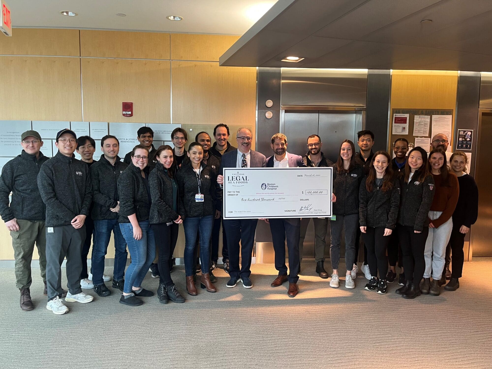 A group of people is standing indoors, smiling and holding a large ceremonial check. They are wearing coordinated black jackets. The setting appears to be a modern office or conference area.