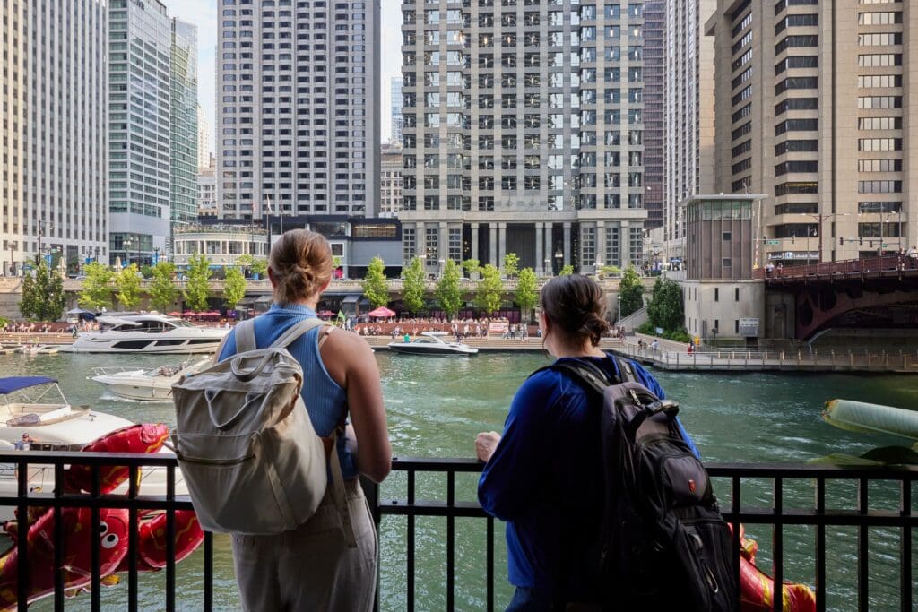 Two people with backpacks stand by a railing, overlooking a river with boats in an urban setting. Tall buildings line the background, and a red bridge is visible to the right. They seem to be enjoying the cityscape on a clear day.